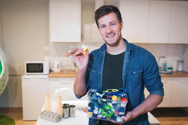 Protéger l'environnement. Couvercles en plastique préparés pour le recyclage. Jeune homme souriant tenant récipient de recyclage rempli de couvercles de bouteilles sur fond de cuisine — Photo