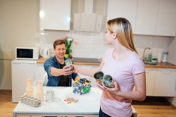 Proteger o ambiente. Jovem sorrindo homem dando a sua namorada latas de metal enquanto ele classifica os resíduos no fundo da cozinha — Fotografia de Stock