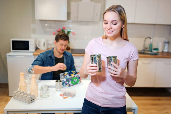 Çevreyi koruyun. Kocası mutfak arka planda atık sıralama yaparken genç gülümseyen kadın yeniden kullanmak için metal teneke kutular tutarak — Stok fotoğraf