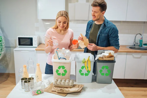 Waste sorting at home. Smiling young couple putting plastic, paper, other waste in garbage bio bins in the kitchen