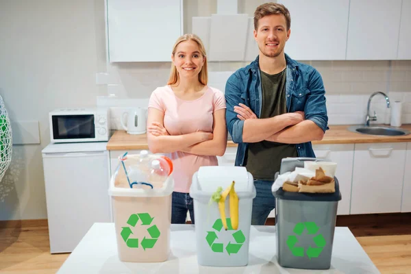 Сортировка отходов дома. Защищать окружающую среду. Молодая счастливая семья выбрасывает отходы в разноцветные мусорные баки с зеленой иконой утилизации на кухне — стоковое фото