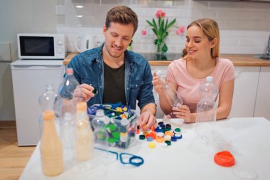Çevreyi koruyun. Evde diğer atık ile masada otururken Aware genç çift boş plastik şişe ve kapaklar geri dönüşüm