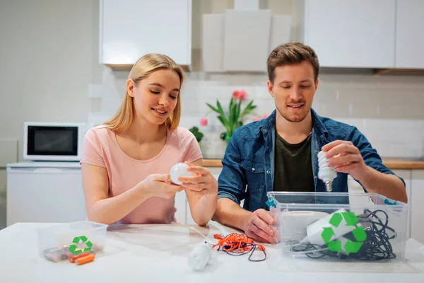 リサイクル、再利用、エネルギー電球、バッテリー、その他の電子廃棄物を、キッチンに座っている間、リサイクルシンボルで容器に選別する若い家族 — ストック写真