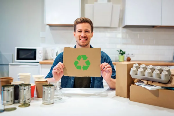 Σύμβολο ανακύκλωσης. Νέος χαμογελαστός άνθρωπος κρατώντας χαρτόνι με εικονίδιο ανακύκλωσης ενώ καθόταν στο τραπέζι με άλλα απόβλητα στο σπίτι — Φωτογραφία Αρχείου