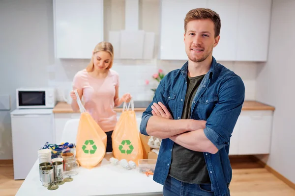 Recycling. junger lächelnder Mann steht nach dem Recycling, während seine Freundin Müllsäcke mit Recycling-Symbol in der Küche hält — Stockfoto