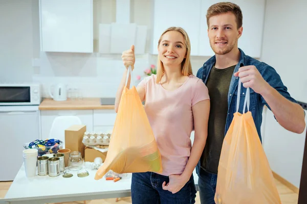 Recycling. verantwortungsbewusste junge lächelnde Familie, die Müllsäcke in der Hand hält, während sie am Tisch steht, der zu Hause mit Abfall gefüllt ist — Stockfoto