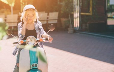 Güzel genç kadın rahat scooter üzerinde oturan ve telefonda konuşurken giyinmiş. Bisiklet üzerinde oturan ve hücresel smartphone kullanarak kız.