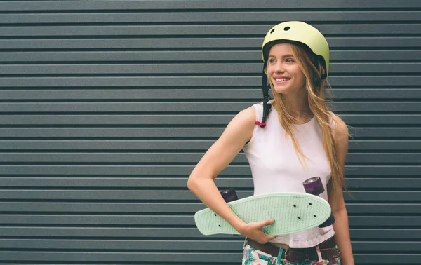 Jovem loira com um skate na mão contra o fundo de uma parede listrada — Fotografia de Stock