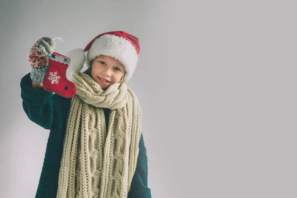小圣诞袜子在手对着女孩。这孩子穿着毛衣, 圣诞帽和围巾工作室拍摄. — 图库照片
