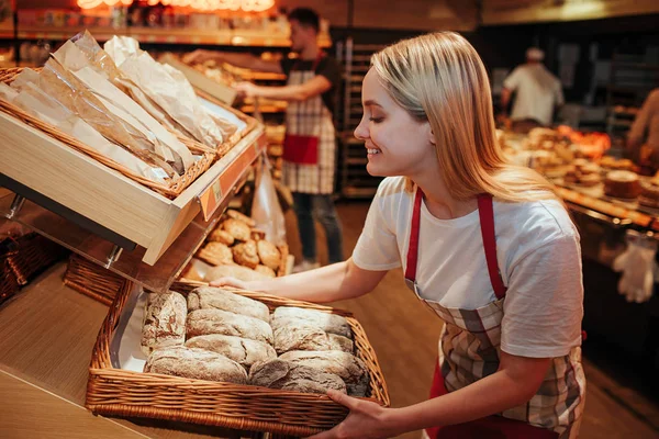 Молодая женщина держит пекарню со свежим хлебом в продуктовом магазине. Она положила его на полку и улыбнулась. Вкусно и опасно. Работаю внутри. Теплый свет . — стоковое фото
