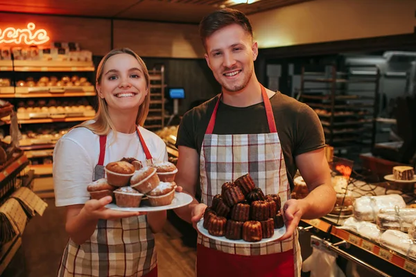 Счастливые молодой человек и женщина стоят в продуктовом магазине или пекарне. Они держат тарелки с блинами и улыбаются. Люди показывают их в камеру. . — стоковое фото