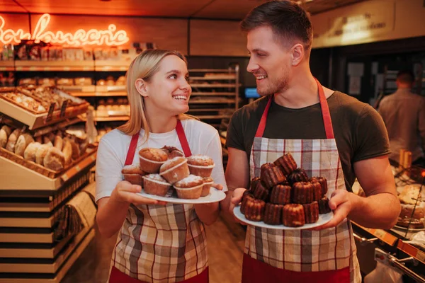 Счастливые молодой человек и женщина стоят в продуктовом магазине или пекарне. Они держат тарелки с маленькими тортами и смотрят друг на друга. Позитивные работники улыбаются . — стоковое фото