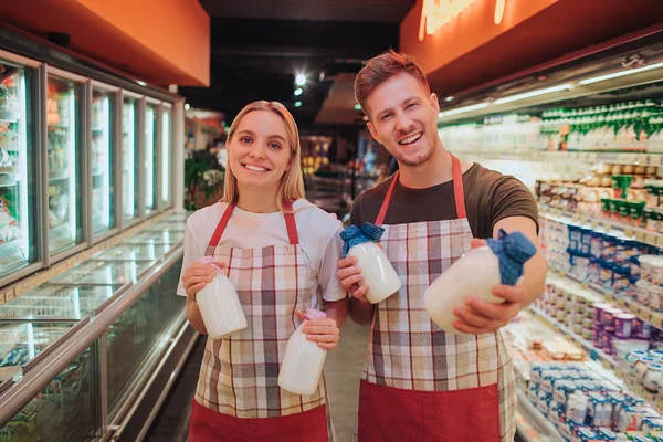 Молодой человек и женщина стоят в продуктовом магазине и молочной полке. Они держат стеклянные бутылки с молоком и позируют перед камерой. Позитивные счастливые рабочие улыбаются . — стоковое фото