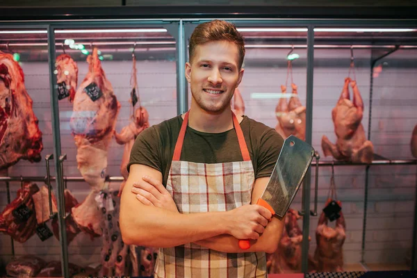 Joven parado en el estante de la carne en la tienda de comestibles. Él sostiene el cuchillo de carnicero en la mano y posar. Detrás de los animales. Sonrisa positiva del trabajador masculino . — Foto de Stock