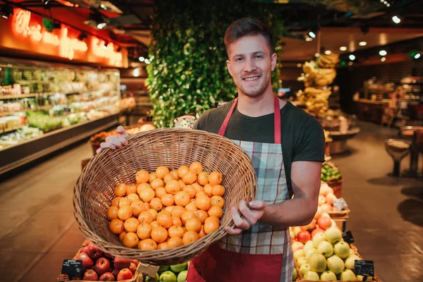 O jovem está em caixas de frutas na mercearia. Ele segura cesta com laranjas e posar na câmera. Sorriso jovem positivo . — Fotografia de Stock
