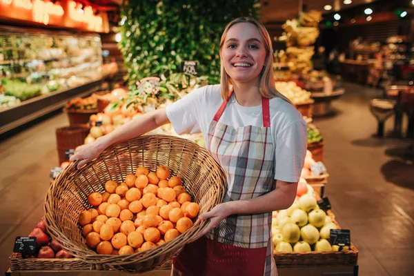 Jovem mulher ficar em caixas de frutas na mercearia. Ela segura cesta com laranjas e posar na câmera. Usa avental. Sorriso de trabalhador positivo. Ela parece feliz. . — Fotografia de Stock