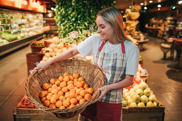 Jovem mulher ficar em caixas de frutas na mercearia. Ela segura o cesto com laranjas e olha para elas. Trabalhador feliz positivo na loja . — Fotografia de Stock