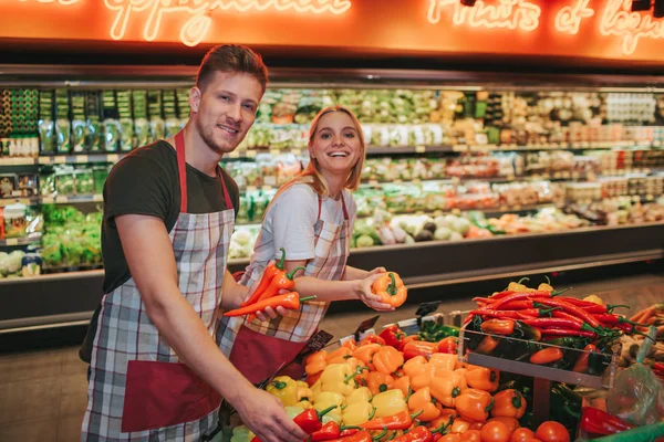 Young man and woman stand at vegetable basket in grocery store. They put some peppers and pose on camera. Positive smiley people.
