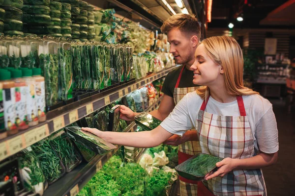 Jovem e mulher estão na prateleira da vegetação na mercearia. Colocam-no cuidadosamente na prateleira e sorriem. Trabalhadores felizes . — Fotografia de Stock