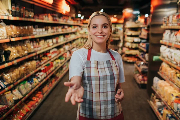 Young woman stand in line between two pasta shelfs in grocery store. She reach hand to camera and smile. Model posing.