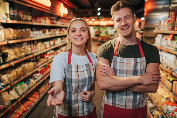 Счастливые молодой человек и женщина стоят в продуктовом магазине среди полки макарон. Они позируют на камеру и улыбаются. Молодая женщина тянется к камере . — стоковое фото