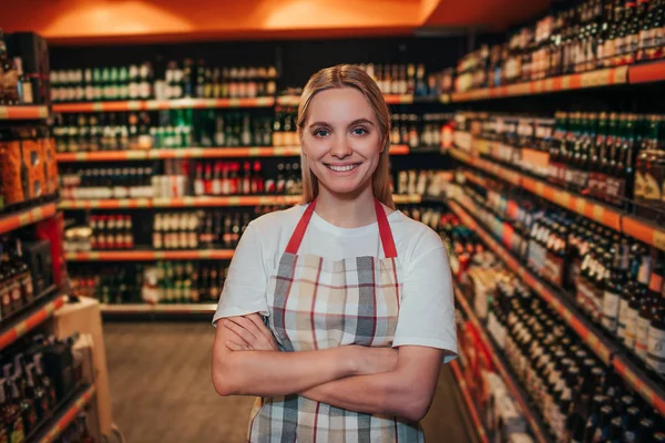 Young woman stand in grocery store among alcohol shelfs. She hold hands crossed and smile. Posing on camera. Wearing apron alone.