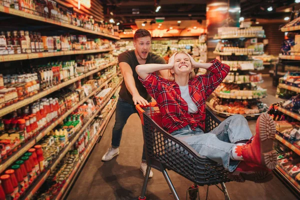 Young couple in grocery store. They play together. Woman sit in trolley and keep eyes closed. Guy scream behind and let trolley go. Fun and happiness.