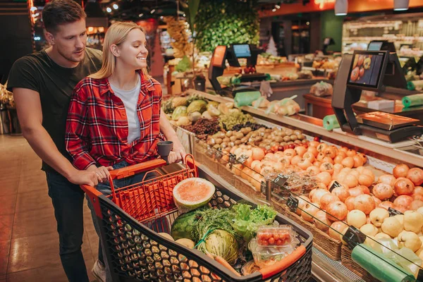 Young couple in grocery store. Positive woman look at pomegranats and other fruit on shelf and smile. Guy help her to carry trolley behind. He look at same direction.