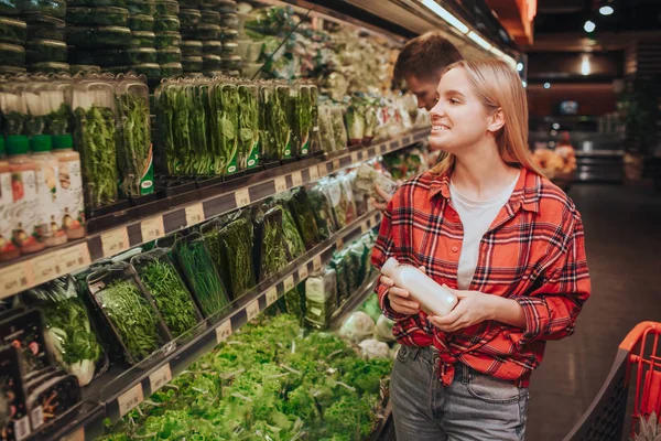 Jovem loira alegre mulher no supermercado. Ela olha para prateleiras com ervas diferentes e sorri. Jovem mulher nake uma escolha. Feliz e positivo . — Fotografia de Stock