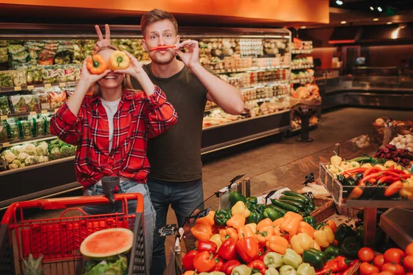 Young couple in grocery store. Funny family pose on camera and smile. She cover eyes with peppers. He hold long pepper instead of moustache and hold two fingers behind her head.