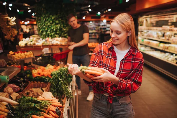 Young couple in grocery store. Woman stand in front and hold carrot in hands. She look at it and smile. Young man stand behind and look at woman. He point on hand.