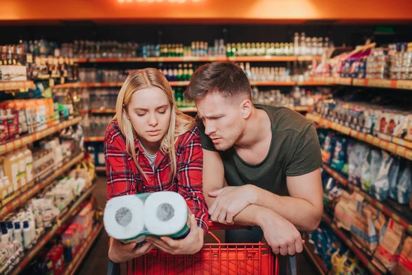 Young couple in grocery store. They lean to troley and look at toilet paper in woman hands. Reading prescription. Concentrated and serious buyers.