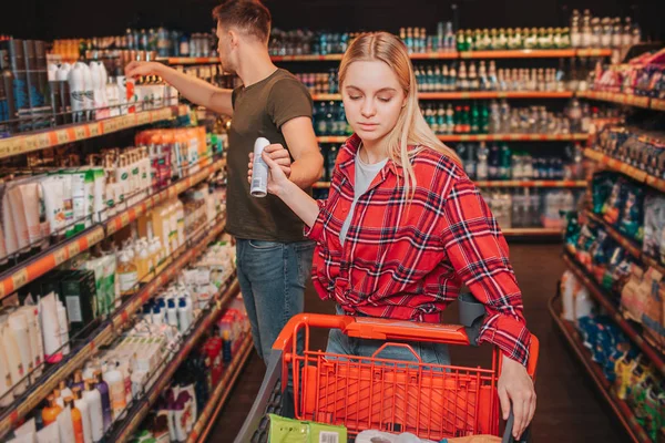 Young couple in grocery store. Woman get deodorant from man. He look at shelf with hygiene goods. She put it into grocery trolley. Serious concentrated buyers.
