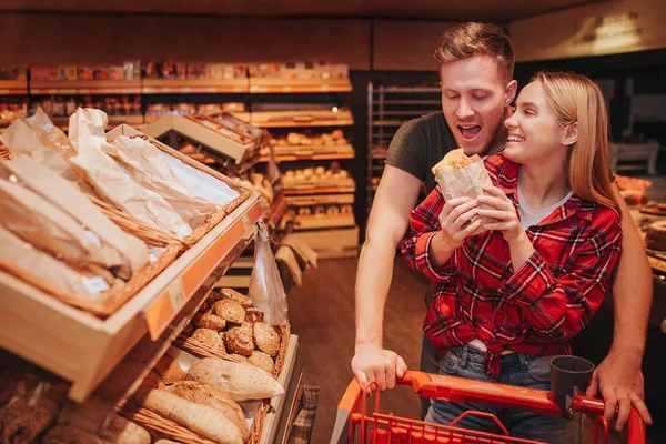 Young couple in grocery store. Funny man hold trolley with hands and want to bite roll. Woman hold it and smile. People stand at bread shelf.