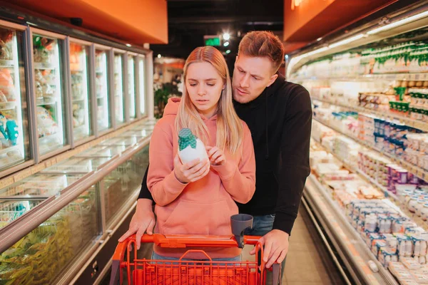 Pareja joven en la tienda de comestibles. La gente cuidadosa lee los ingredientes en la botella de leche. Serio y concentrado. Estantería de yogur . —  Fotos de Stock
