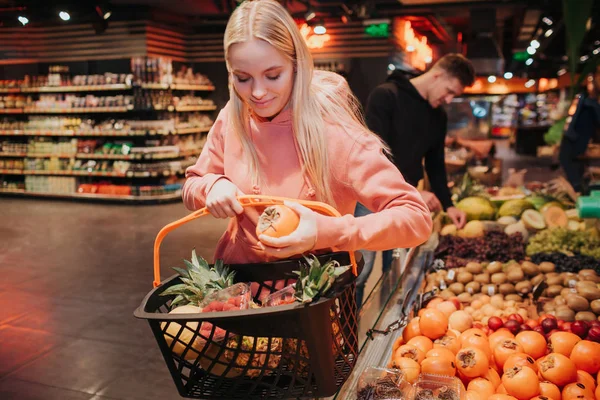 Casal jovem na mercearia. Uma mulher simpática colocou cáqui no cesto do supermercado e olhou para baixo. O tipo fica atrás. Ele apanha fruta. . — Fotografia de Stock