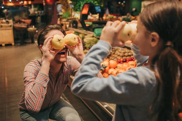 Jovens pais e filhas na mercearia. Eles brincam com maçãs. As pessoas escondem eues com fruta. Família divertida . — Fotografia de Stock