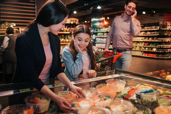 Junge Eltern und Tochter im Lebensmittelgeschäft. Frau hält Schachtel mit Salat in den Händen und lächelt. Mädchen stehen daneben und betrachten es. Junger Mann telefoniert und schaut sich Modelle an. — Stockfoto