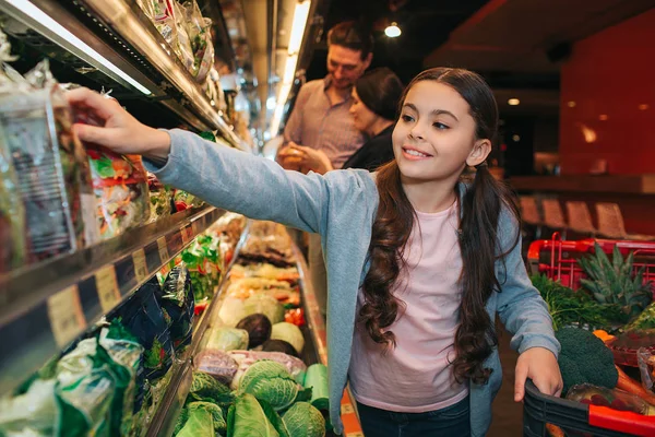 Junge Eltern und Tochter im Lebensmittelgeschäft. kleines Mädchen nimmt Salat und lächelt. sie reicht die Hand zum Regal. Eltern stehen dahinter. — Stockfoto