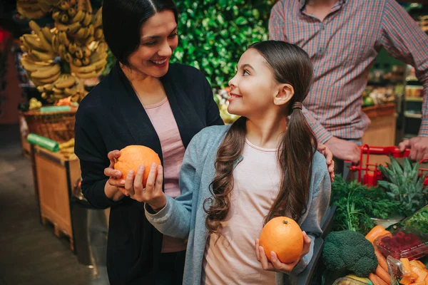 Молодые родители и дочь в продуктовом магазине. Девочка смотрит на родителей и улыбается. Она держит апельсины в руках. Отец, встань позади . — стоковое фото
