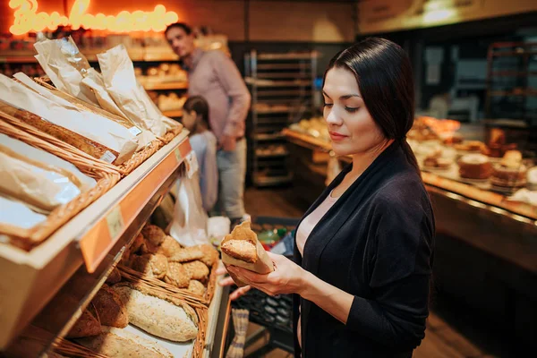 Jovens pais e filha na mercearia. Mulher ficar na frente e segurar o pão nas mãos. Ela olha para isso seriamente. Pai e filha ficam atrás , — Fotografia de Stock