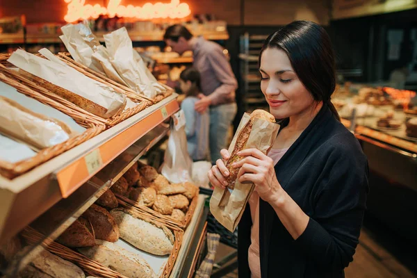 Молодые родители и дочь в продуктовом магазине. Веселая женщина чувствует запах хлеба, который держит в руках. Отец стоит позади с дочерью и несет тележку с продуктами. . — стоковое фото