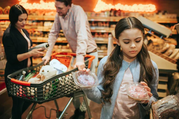 Junge Eltern und Tochter im Lebensmittelgeschäft. Mädchen halten zwei Schachteln mit Donuts und betrachten sie. Eltern stehen am Einkaufswagen. — Stockfoto