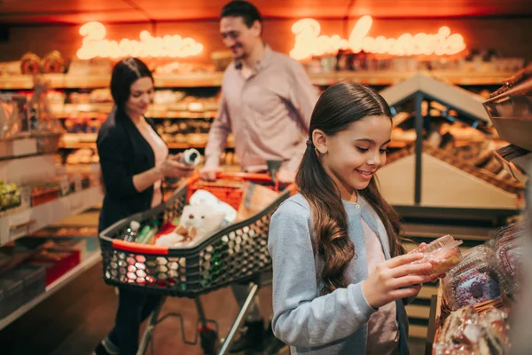 Jovens pais e filha na mercearia. alegre pequena menina segurar caixa com donut e sorriso. Os pais ficam atrás no carrinho com mercearia . — Fotografia de Stock