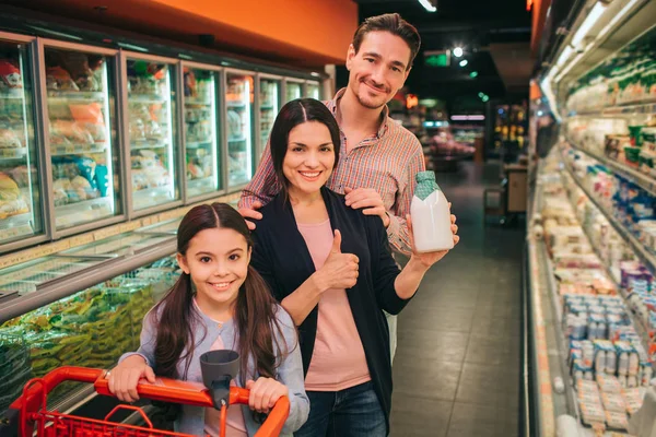 Junge Eltern und Tochter im Lebensmittelgeschäft. Sie stehen zwischen Produktregalen und posieren vor der Kamera. Mädchen halten Trolley. Gruppenlächeln. — Stockfoto