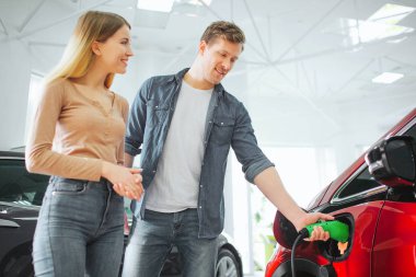 Showroom ilk elektrikli araba satın genç gülümseyen çift. Adam güç kablosu kaynağı ile modern çevre dostu araç şarj takılı. Otomobilin geleceği