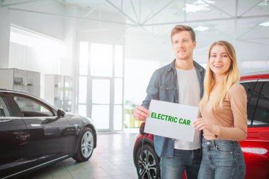 Genç aile showroom ilk elektrikli araba satın. Çevre kırmızı araç yakınında ayakta iken kelime elektrikli araba ile kağıt tutarak çekici çift gülümseyen. Elektrikli araba satış konsepti