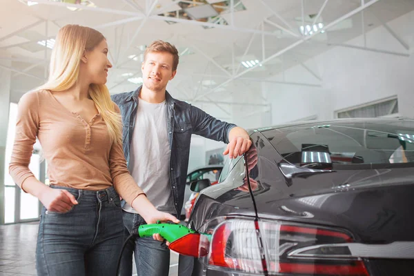 Giovane coppia di famiglia sorridente acquistare prima auto elettrica nello showroom. Primo piano di donna attraente che carica auto ibrida ecologica con l'alimentazione del cavo collegato mentre guarda suo marito — Foto Stock