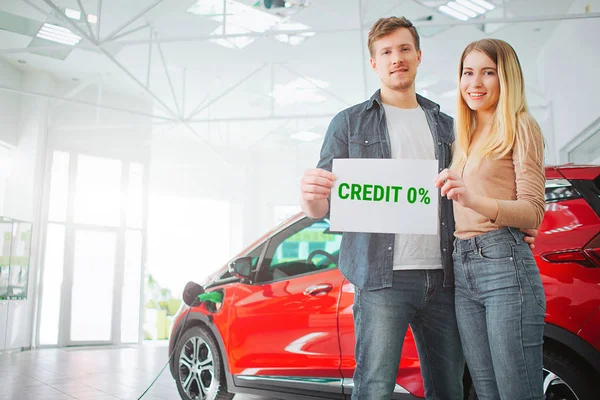 La giovane famiglia acquista la prima auto elettrica nello showroom. Sorridente attraente coppia in possesso di carta con la parola di credito mentre in piedi vicino eco veicolo rosso. Batteria auto elettrica per l'ecologia . — Foto Stock