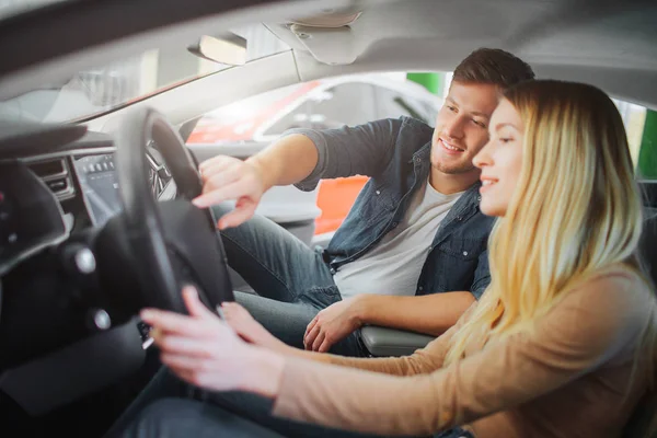 Joven familia comprando el primer coche eléctrico en la sala de exposición. Atractiva pareja sonriente mirando los botones en el volante en un vehículo ecológico de lujo antes de probar la conducción. Concepto de venta de coches eléctricos — Foto de Stock